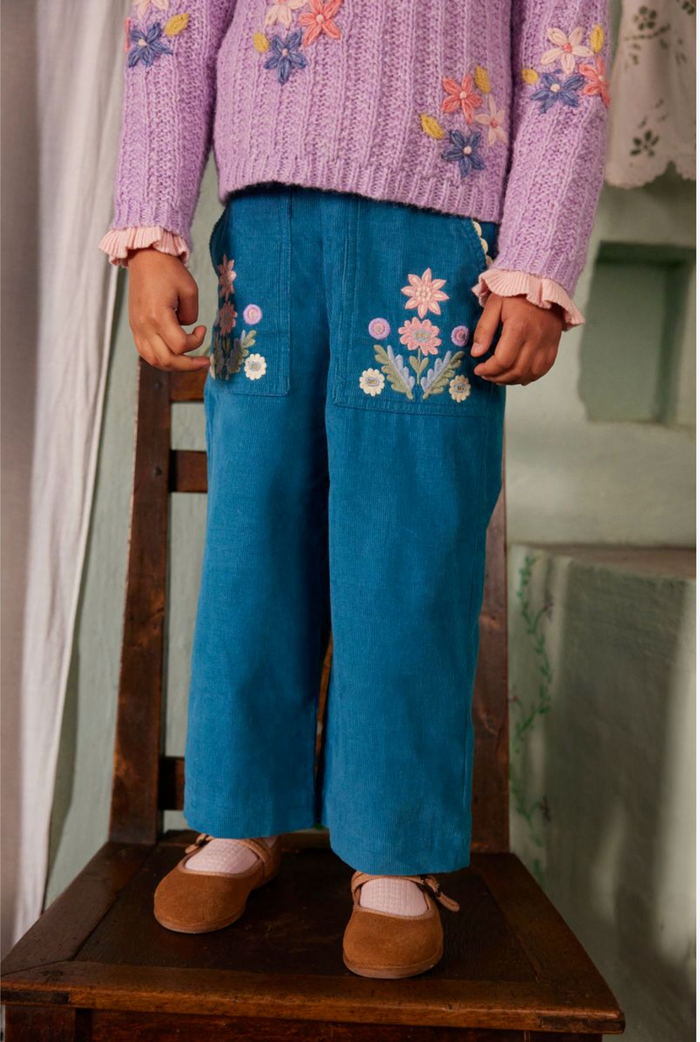 A child wearing a lavender knit sweater and blue wide-leg trousers with colorful floral embroidery on the pockets, sitting on a wooden chair.