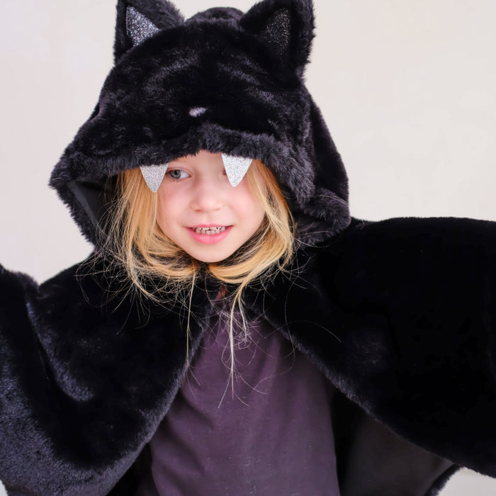 A young girl wearing a black plush bat costume cape with attached hood, cat ears, and silver glitter bat wings peeking out from under the hood, photographed against a plain light background.