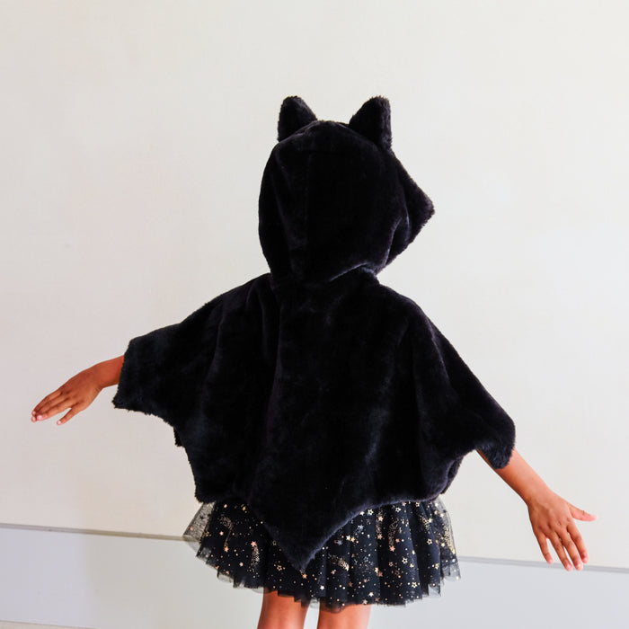 A child wearing a black plush bat cape with attached ears and a sparkly black tulle skirt, photographed from the back against a plain light wall.