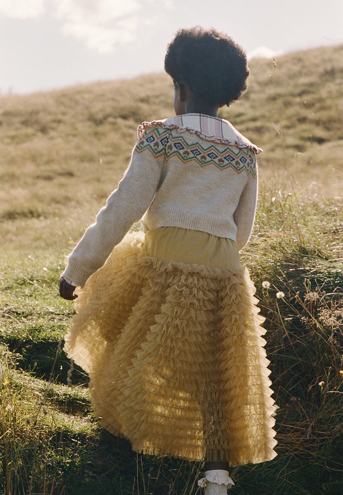 Person wearing a patterned cardigan and yellow skirt standing in a grassy field.