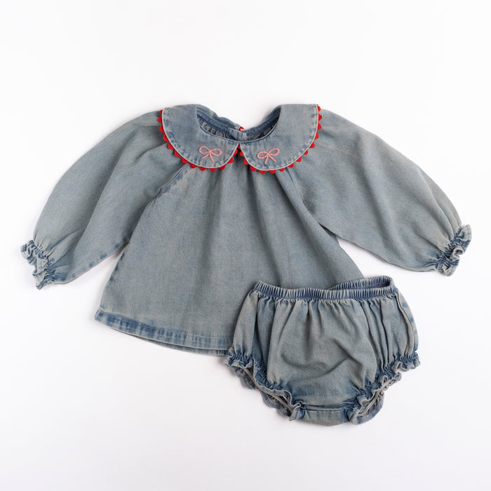 A baby girl's two-piece outfit made of washed denim, featuring a long-sleeve top with a Peter Pan collar trimmed in red and pink embroidery, and matching ruffled bloomers, laid flat on a white background.