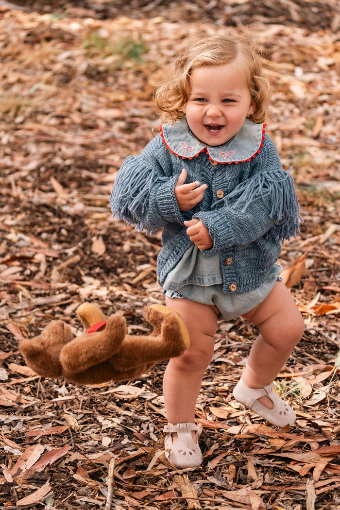 A toddler girl wearing a blue fringed knit cardigan with a patterned collar and denim shorts, smiling and running outdoors on a bed of dry brown leaves. A brown plush toy lies on the ground nearby.