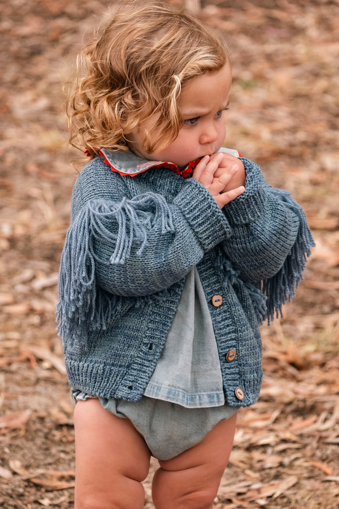 A toddler standing outdoors on a leaf-covered ground, wearing a blue fringed knit cardigan over a light blue denim shirt and gray diaper.