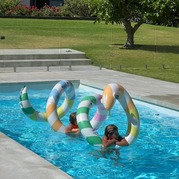 Two children playing with colorful inflatable giant snake n a pool surrounded by grass and a tree.