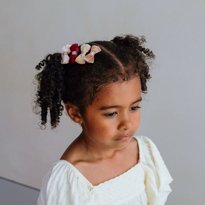 A young child with curly dark hair styled in two pigtails, wearing a cluster of fabric hair bows in red, pink, and white, paired with a white top against a plain light gray background.