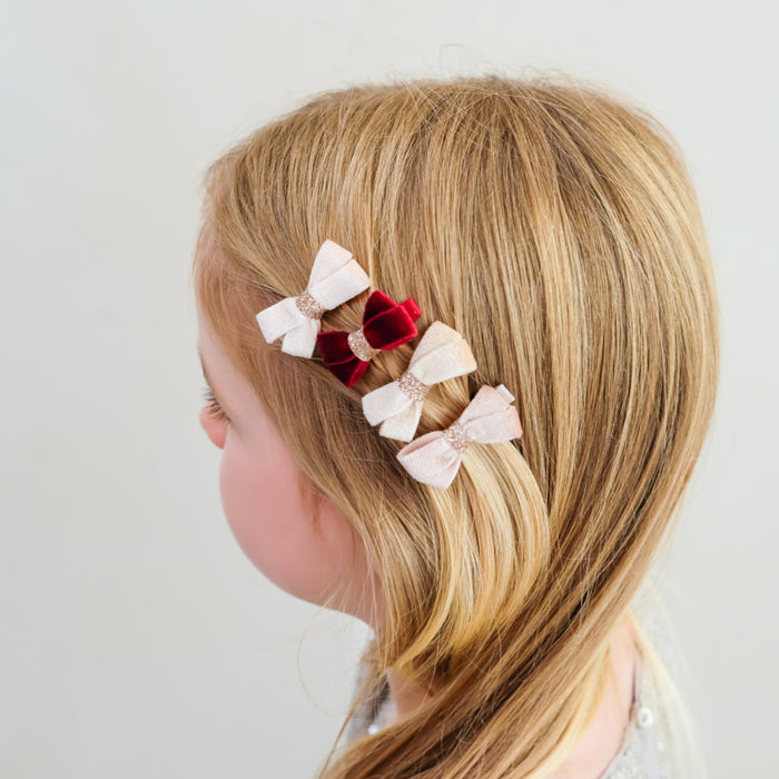 small bow hair clips in pale pink, deep red velvet, and light beige with glitter trim, shown attached to a child's blonde hair against a plain light background.