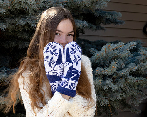Woman wearing the Gnomes in Love mittens in navy and white stranded colorwork featuring gnome motifs, posed in front of a blue spruce tree
