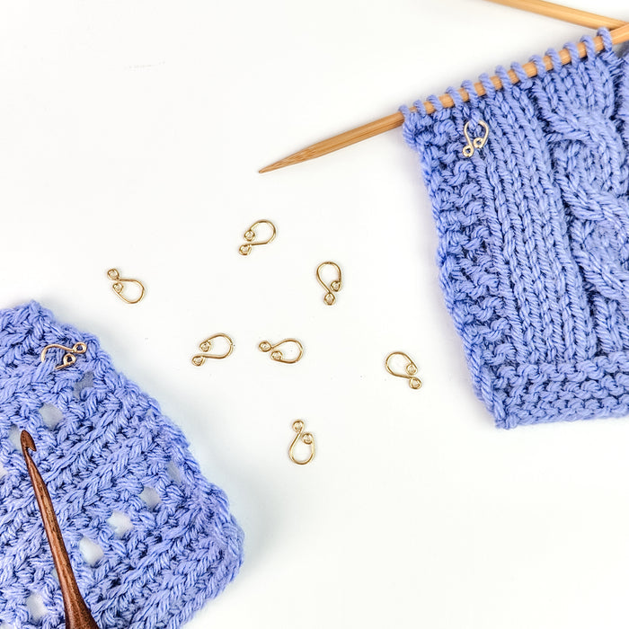 Gold cast melody clip stitch markers scattered between blue crochet and knit swatches with hooks and needles