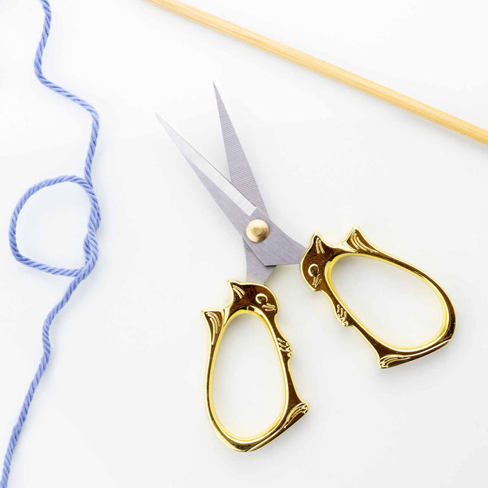 Golden cat embroidery scissors open with blue yarn and a bamboo knitting needle showing the sharp pointed blades