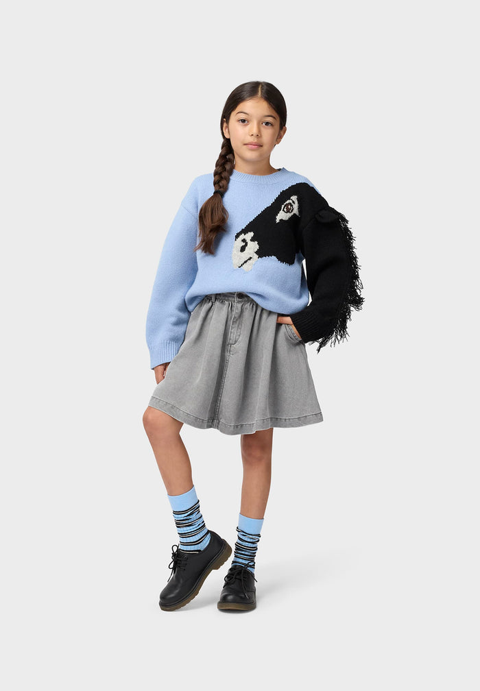 Young girl wearing a light blue sweater with a black and white design, gray skirt, and black shoes on a white background.