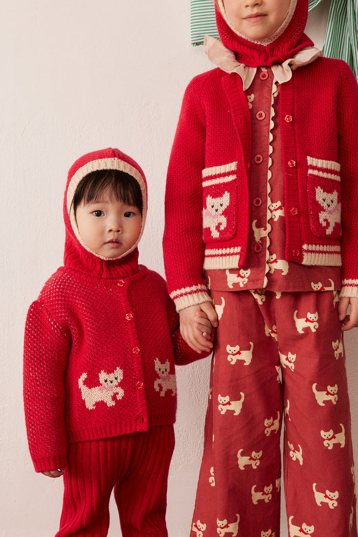 Two children wearing matching red knit holiday outfits with cream dog motifs. The child on the left wears a red cardigan and hooded sweater, while the taller child on the right wears a matching red cardigan, hood, and wide-leg pants.