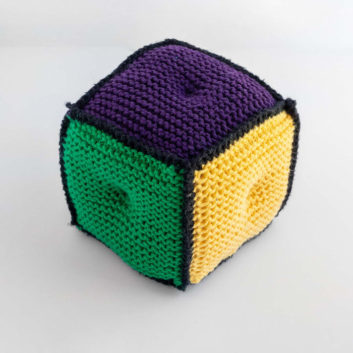 Close-up of a knitted game die with purple top, green side, and yellow side, all edged in black yarn