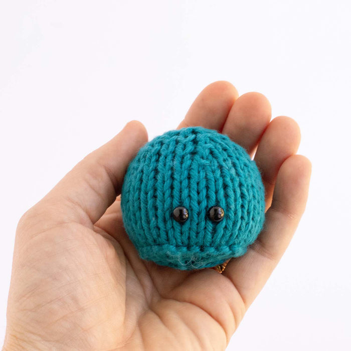 Hand holding a miniature teal knitted gumdrop game piece with black bead eyes