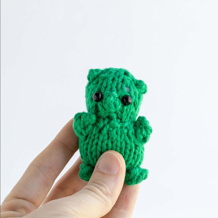 Green knitted gummy bear toy with black bead eyes centered among scattered colorful real gummy bear candies in orange, red, yellow, and green on white background