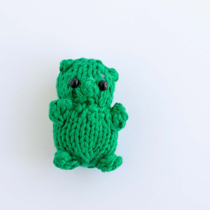 Fingers holding a tiny green knitted gummy bear toy with black bead eyes, small rounded ears, and outstretched arms to show its miniature scale