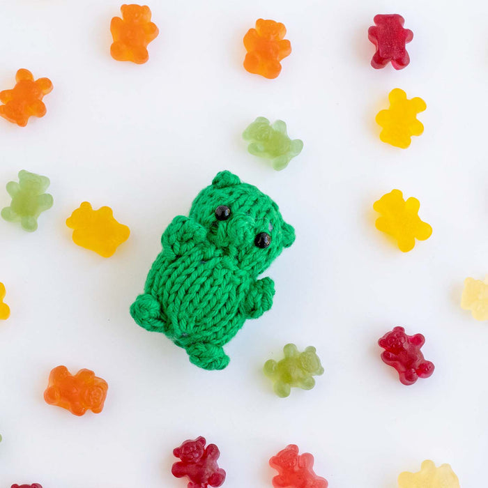 Green knitted gummy bear toy lying flat on white background showing full body with black bead eyes, small ears, round belly, and stubby arms and legs