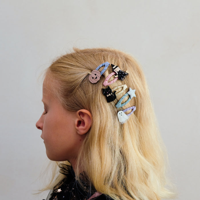 A side-profile view of a young girl with blonde hair wearing a set of colorful, whimsical hair clips, including a pumpkin, a ghost, a star, and a small cat, against a plain light gray background.
