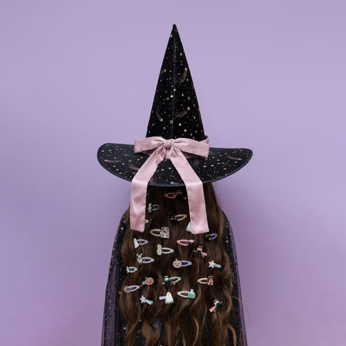 A person wearing a black witch hat with a pink ribbon bow and a matching black cape, with long brown hair adorned with many colorful hair clips, set against a solid lavender background.