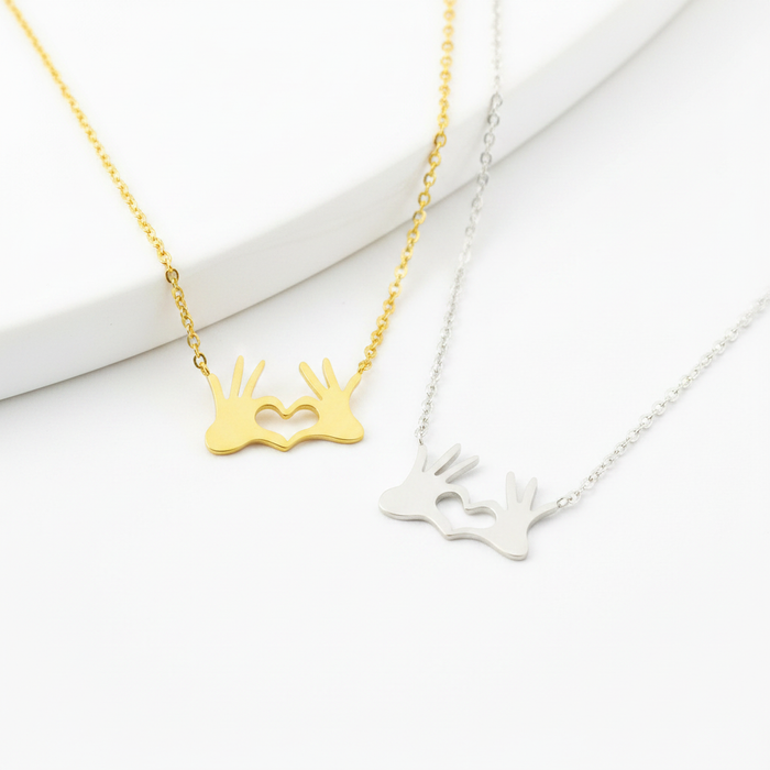Two delicate necklaces featuring a pendant of two hands coming together to form a heart shape with the fingers. One necklace is finished in polished gold and the other in silver. Both pendants are integrated into fine cable chains and displayed on a clean, bright white surface.