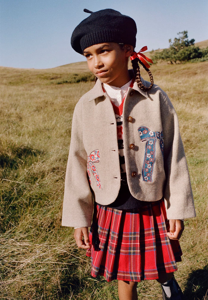 Person wearing a beige jacket with floral patterns and a red plaid skirt in an outdoor setting.