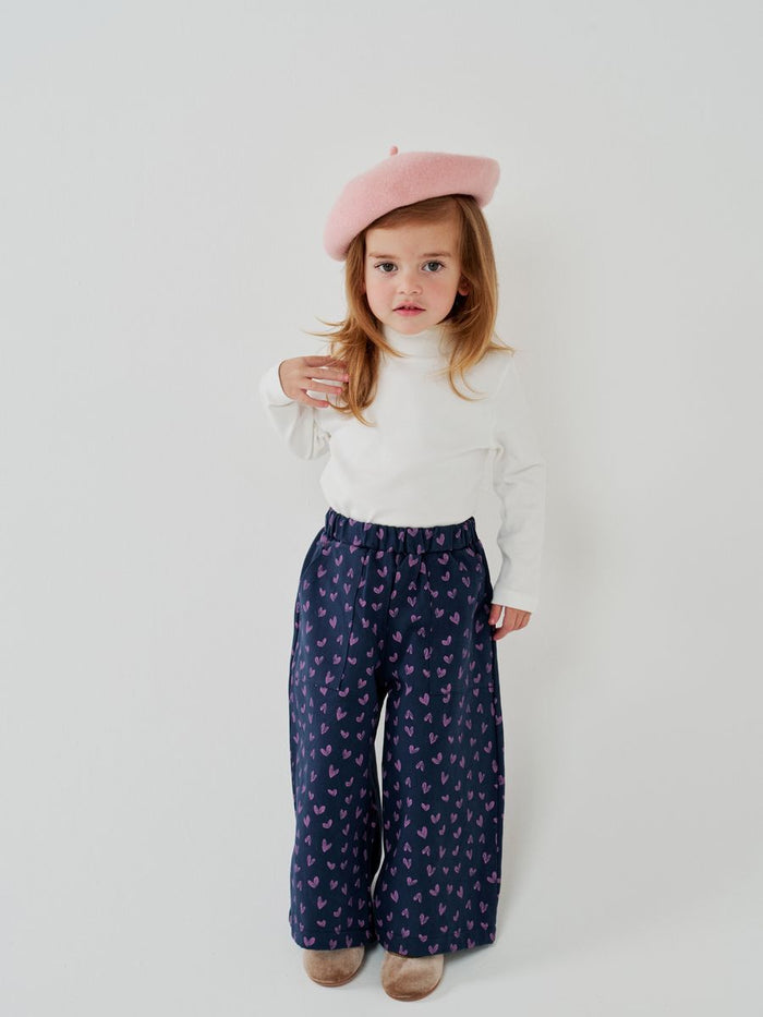 Child wearing a white top, navy pants with pink patterns, and a pink beret on a white background