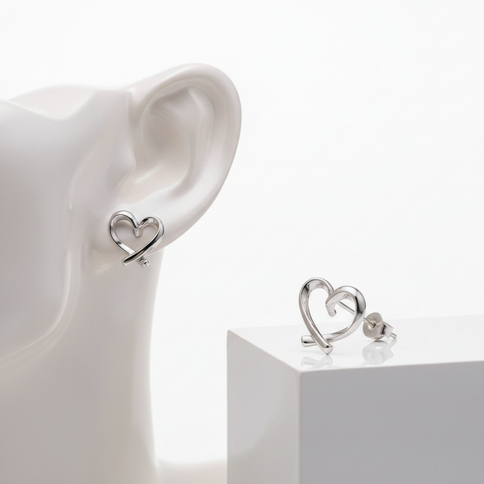 A pair of silver-toned stud earrings featuring a minimalist, open-heart design created from a single flowing ribbon-like strand of metal that overlaps at the bottom. One earring is shown worn on a white mannequin ear, while the other sits on a white rectangular display block, highlighting the butterfly clasp backing. The setting is a clean, all-white studio background.
