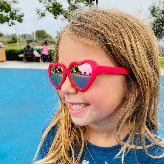 Child wearing heart-shaped sunglasses by a pool with people in the background