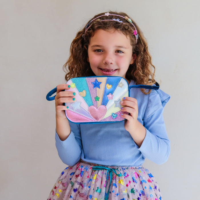 A young girl with curly brown hair wearing a light blue ribbed long-sleeve top and a colorful patterned skirt, holding up a small zippered pouch decorated with a colorful fairy design.