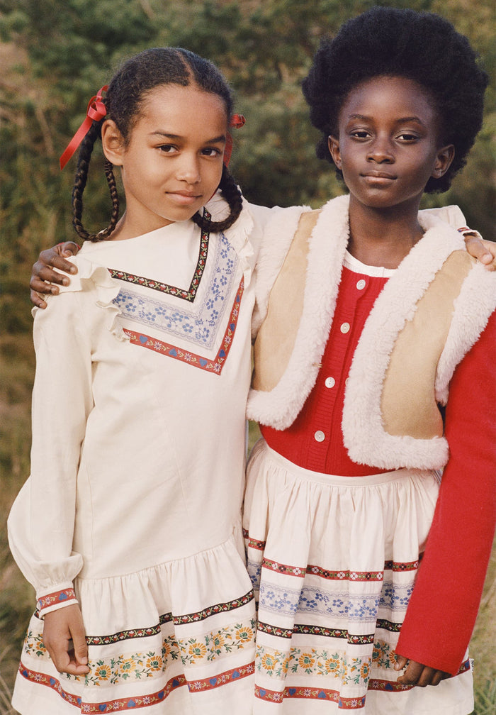 Two young girls in traditional outfits standing outdoors with a natural background.
