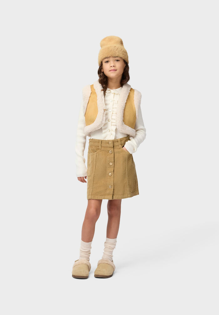 Child wearing a beige vest, skirt, and hat with white sweater on a white background