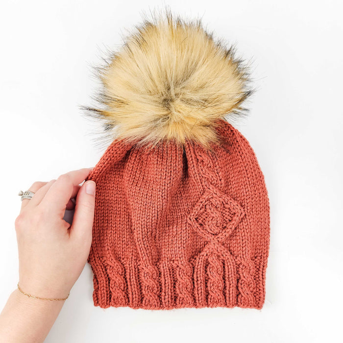 Terracotta cable-knit Holly Beanie with faux fur pom-pom held by hand on white background