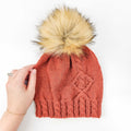 Terracotta cable-knit Holly Beanie with faux fur pom-pom held by hand on white background