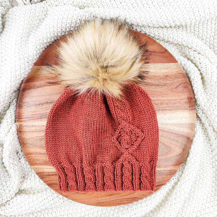 Terracotta cable-knit Holly Beanie with faux fur pom-pom on wooden board with cream knit blanket