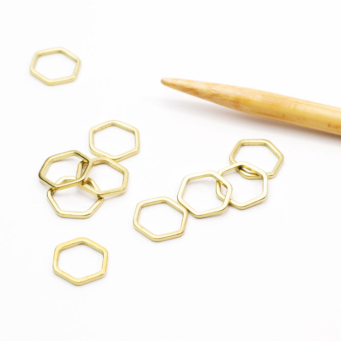 Set of gold hexagonal honeycomb-shaped simple stitch markers scattered on a white surface next to a bamboo knitting needle