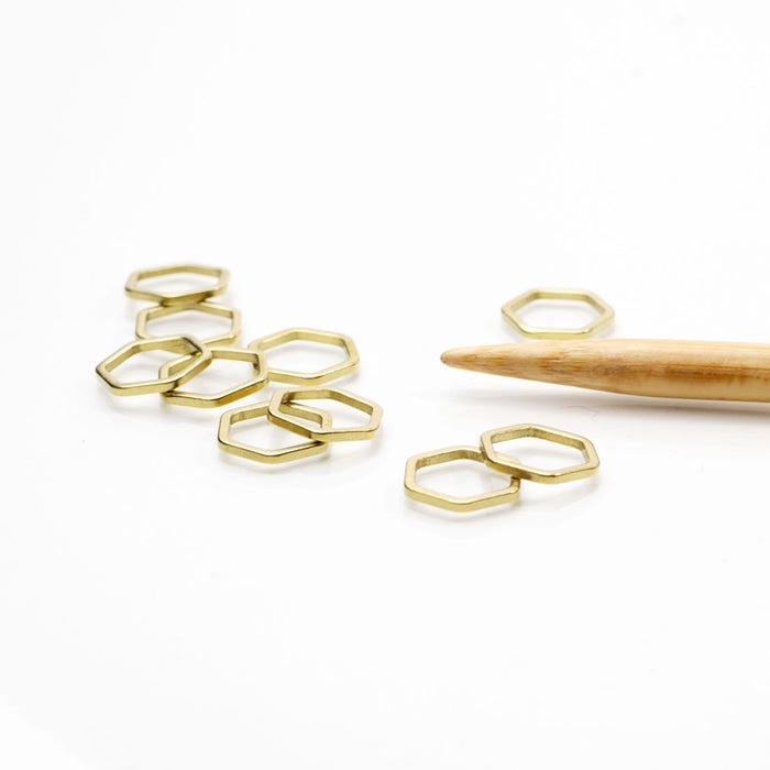 Gold hexagonal honeycomb stitch markers casually grouped on a white surface with a bamboo knitting needle tip showing two size variations