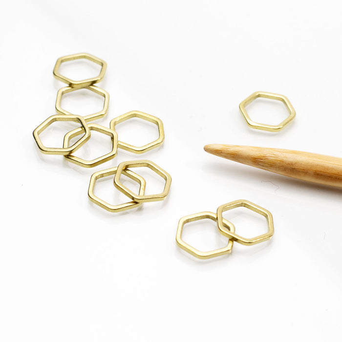 Gold hexagonal honeycomb stitch markers arranged on a white surface with a bamboo knitting needle showing the larger size markers