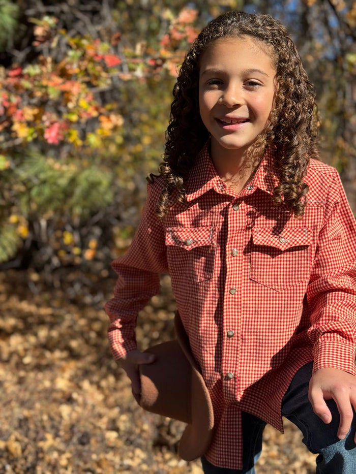 Child in a red checkered shirt holding a cowboy hat outdoors with autumn foliage.