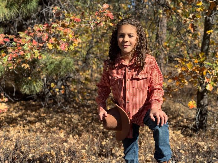 Child in a red checkered shirt and blue jeans holding a cowboy hat in an autumn forest.