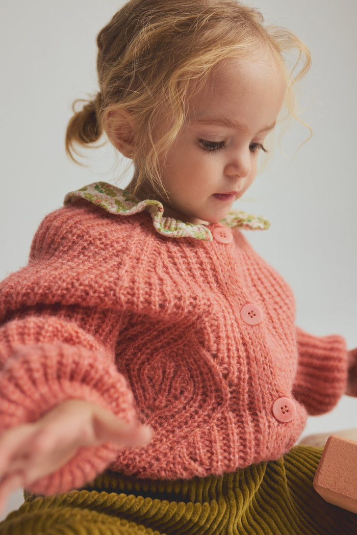 child wearing pink cardigan over collared shirt with corduroy pants on light grey background