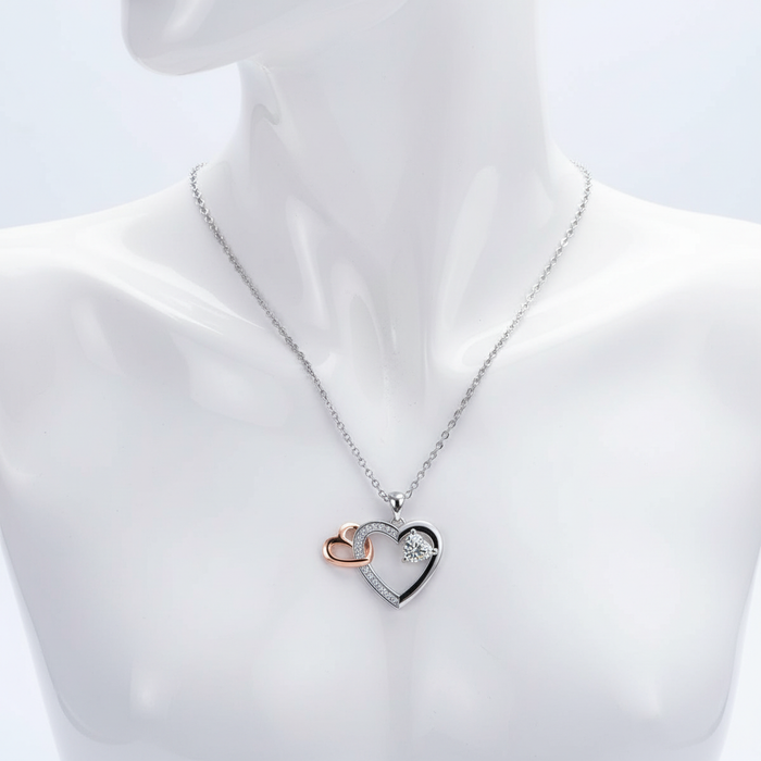 A silver-toned necklace featuring a complex triple-heart pendant displayed on a white mannequin. The design includes a large silver heart partially pavé-set with shimmering crystals, an interlocking small rose gold-toned heart, and a sparkling heart-shaped crystal suspended within the main frame. The pendant hangs from a fine silver-toned cable chain.