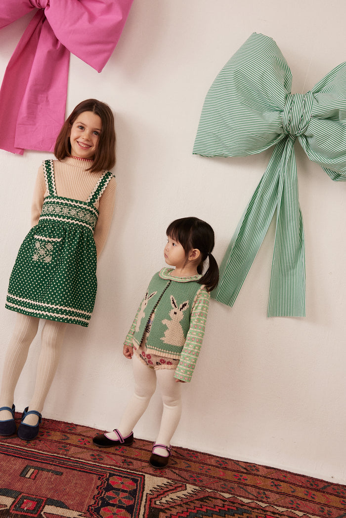 Two young girls modeling knit holiday-themed clothing in a styled indoor setting with large decorative bows on the wall and a patterned rug on the floor. The older girl wears a green polka dot dress, and the younger girl wears a green knit cardigan with a bunny design.