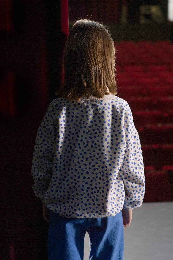 A child viewed from behind wearing a white long-sleeve top with a blue polka dot pattern and blue pants, standing in front of red theater-style seating.