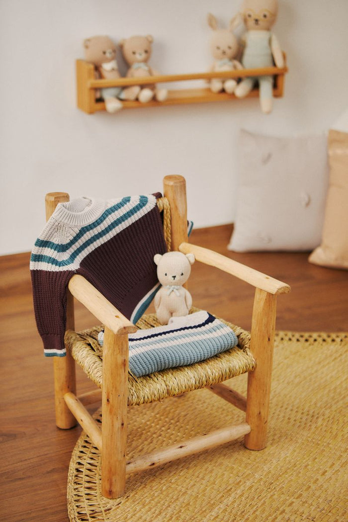 Wooden chair with a blanket and stuffed toy on a wooden floor, shelf with more toys in the background.