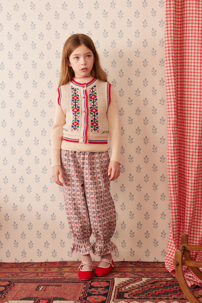 A young girl stands in a vintage-style room wearing a cream knit vest with colorful floral embroidery, matching patterned trousers, and red shoes. The background features patterned wallpaper, a red-and-white checkered curtain, and a patterned rug.