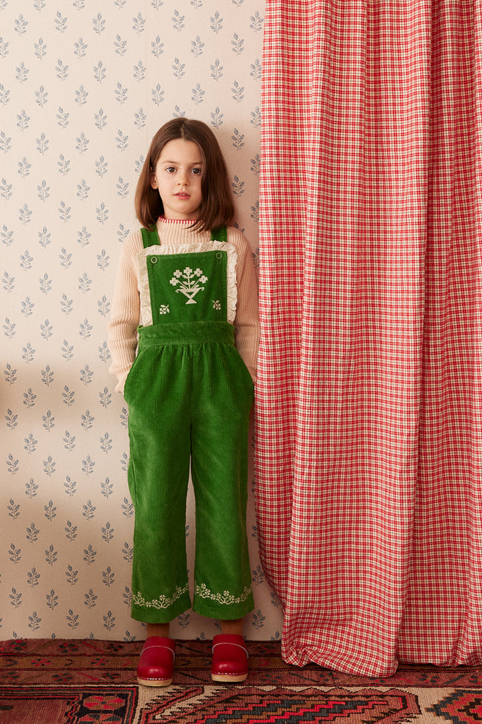 A young girl stands indoors wearing a green corduroy pinafore-style overall with embroidered details, a cream knit top, and red shoes. She is posed beside a red-and-white gingham curtain against patterned wallpaper and a rug.