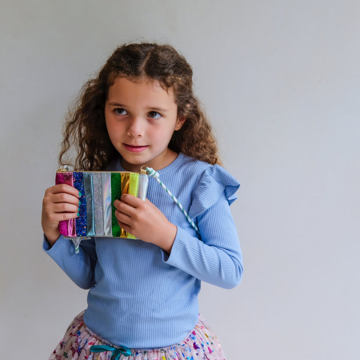 A young girl with curly brown hair wearing a light blue ruffled long-sleeve top and a colorful patterned skirt, holding a small rainbow-striped purse against a plain light gray background.