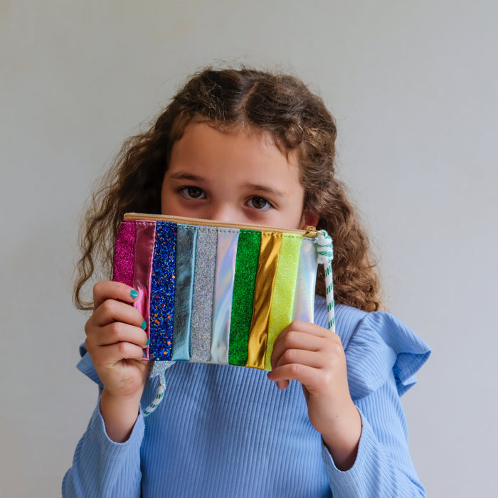 A young girl with curly brown hair wearing a light blue ribbed shirt holds a colorful striped purse in front of her face. The pouch features vertical stripes of metallic pink, glittery blue, silver, green, yellow, and white, with a light blue and white striped zipper pull.