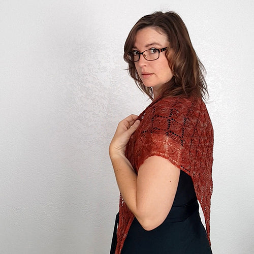 Side profile of model looking over her shoulder wearing the Kellerwald lace shawl in rust red draped across her upper back, paired with a black dress