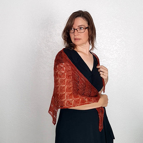 Front view of model holding the Kellerwald lace shawl in rust red wrapped across her chest, showing the open diamond lace pattern detail