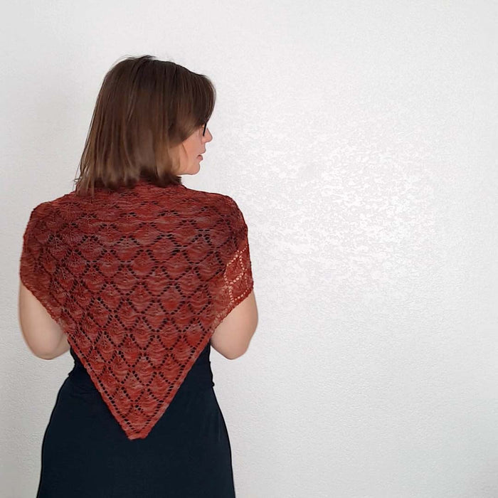 Back view of model wearing the Kellerwald shawl in rust red, showing the full triangular shape and repeating diamond lace motif from behind